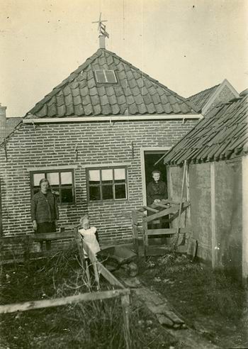 Achterkant winkel/woonhuis Vrouw Helder, die rechts in de deur staat. Links een dochter, op het tuinpad een kleinkind. 