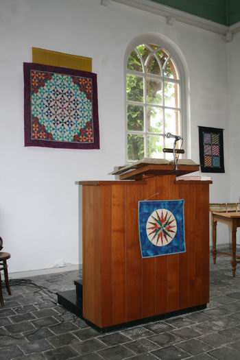 Resize of OM (14) 12092009 Het spreekgestoelte in de vermaning, omlijst door kleurige quilts; het licht laat zich gelden