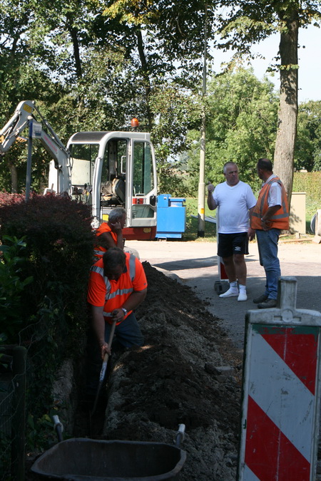 Dicht tegen de haag van de tuin van de familie Kuiper zijn de kabelaars bezig sleuven te graven, kabels te leggen cq. vervangen, sleuven weer dicht te gooien.... Bewoner Ties Kuiper (wit shirt) in gesprek met de machinebediener. Twee specialisten op de schop voor het echte handwerk....